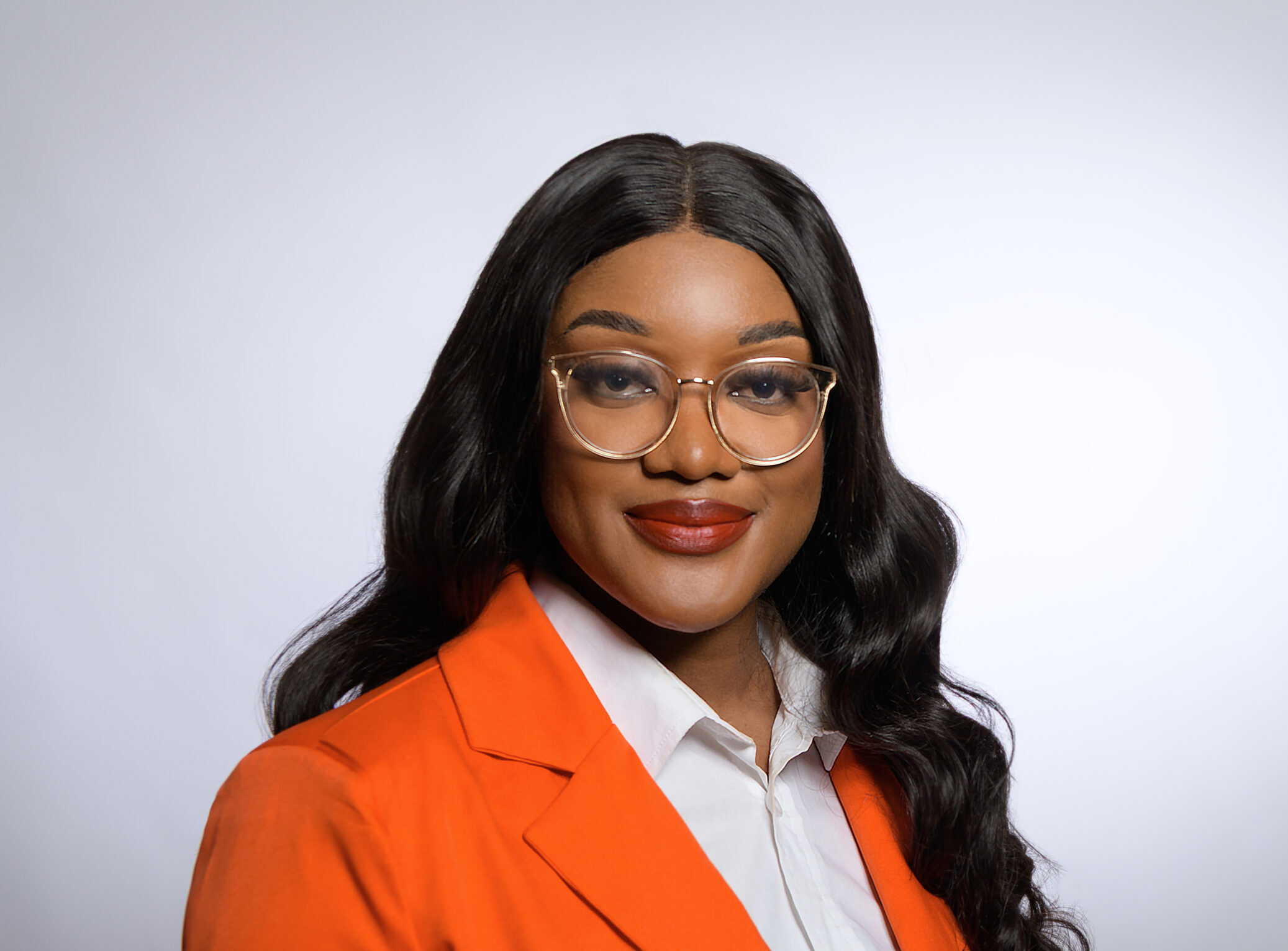 Professional headshot of a smiling woman with long black hair, wearing glasses and an orange blazer over a white shirt.