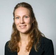 Professional head-and-shoulders portrait of a woman with wavy light brown hair, wearing a dark blazer, smiling against a light gray background.