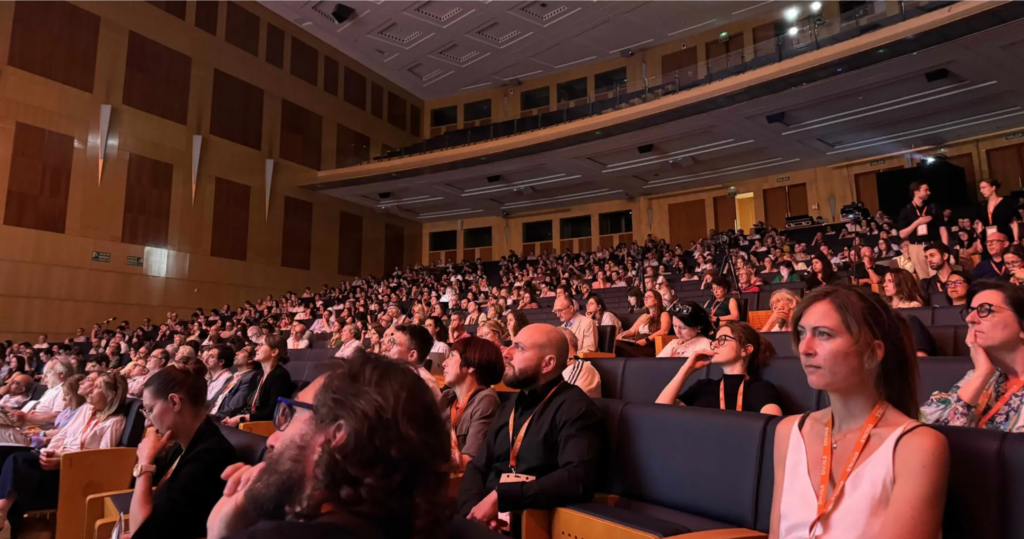 APST_Newsletter 2026_Ambulanzpartner Forschung beim ENCALS Meeting_Bild 2_20260708 Large auditorium packed with attendees seated in rows, many wearing lanyards, watching a presentation at a conference.