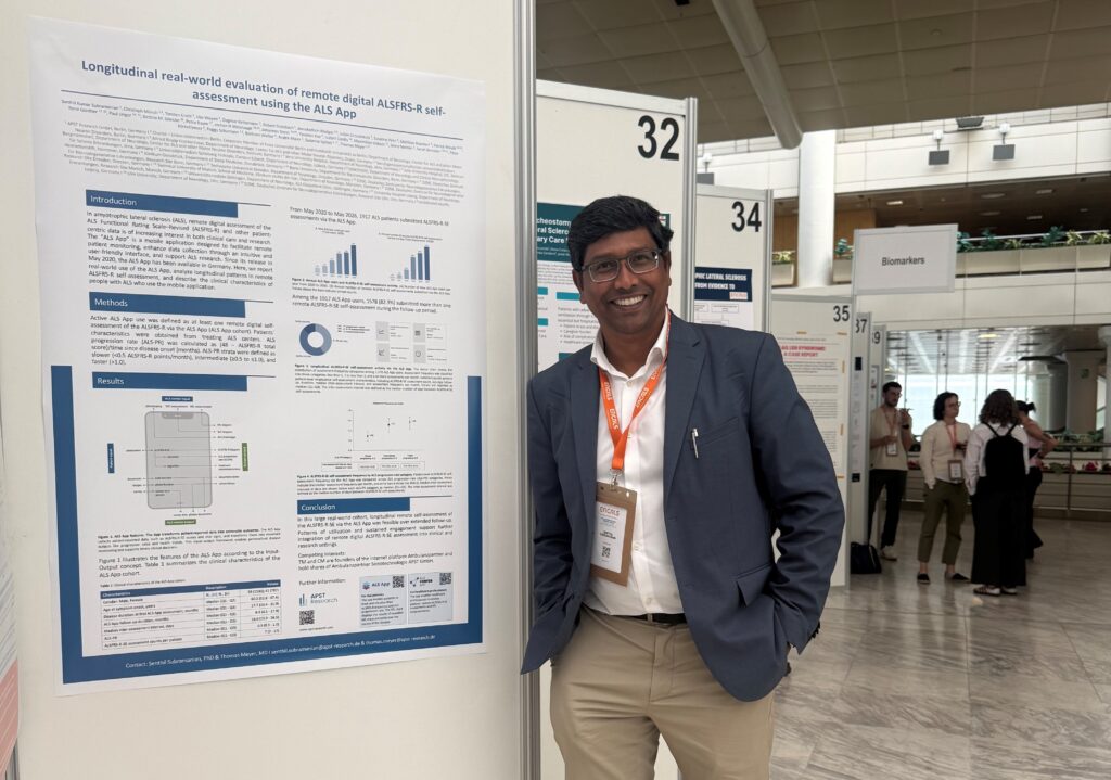 APST_Newsletter 2026_Ambulanzpartner Forschung beim ENCALS Meeting_Bild 520260708 Smiling man in a suit stands beside a research poster at a conference, booth 32, with other attendees in the background.